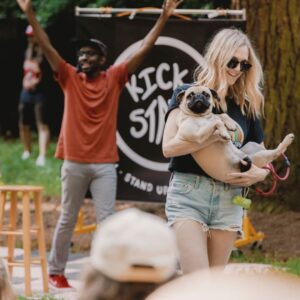 A man and a woman holding a dog in front of a banner that says Kickstand Comedy in the Park